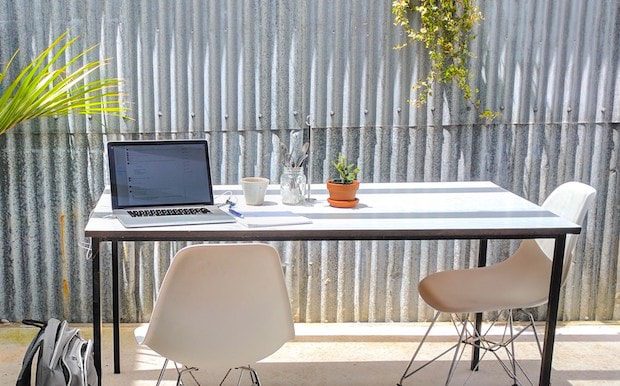 image of a writers desk while working on an eBook. Outside, laptop to the left side, one chair to the right of the table, and one immediately in front of the laptop. Palm trees in front of fence serve as the backdrop.
