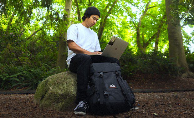 Man writing on his Substack Publication while sitting on a rock in the forest, on his laptop with a black suitcase.