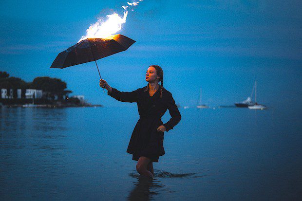 woman standing in dark water against a dark sky, holding a black umbrella on fire exemplifying the severity of some decisions